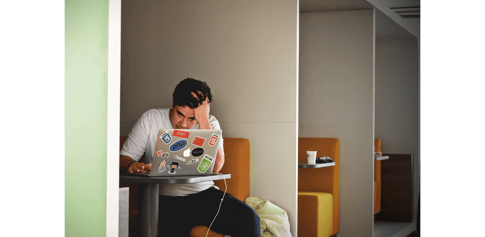 A man in a white t-shirt sitting in a booth while struggling with something on his laptop